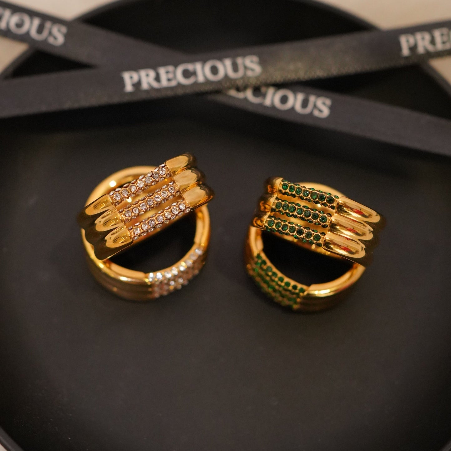 Gold earrings with gemstones on a black dish, with 'PRECIOUS' branding visible.