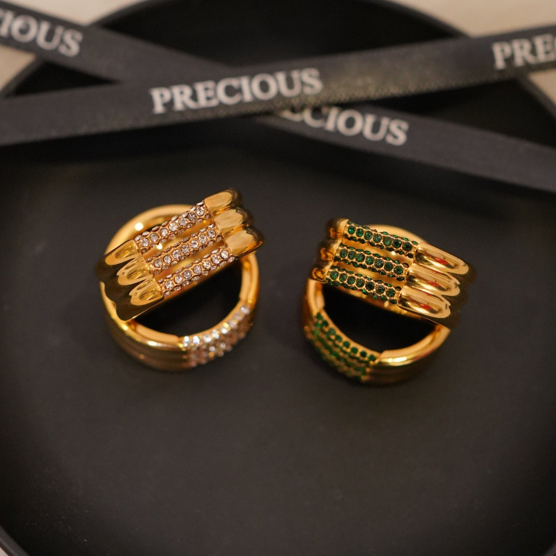Gold earrings with gemstones on a black dish, with 'PRECIOUS' branding visible.